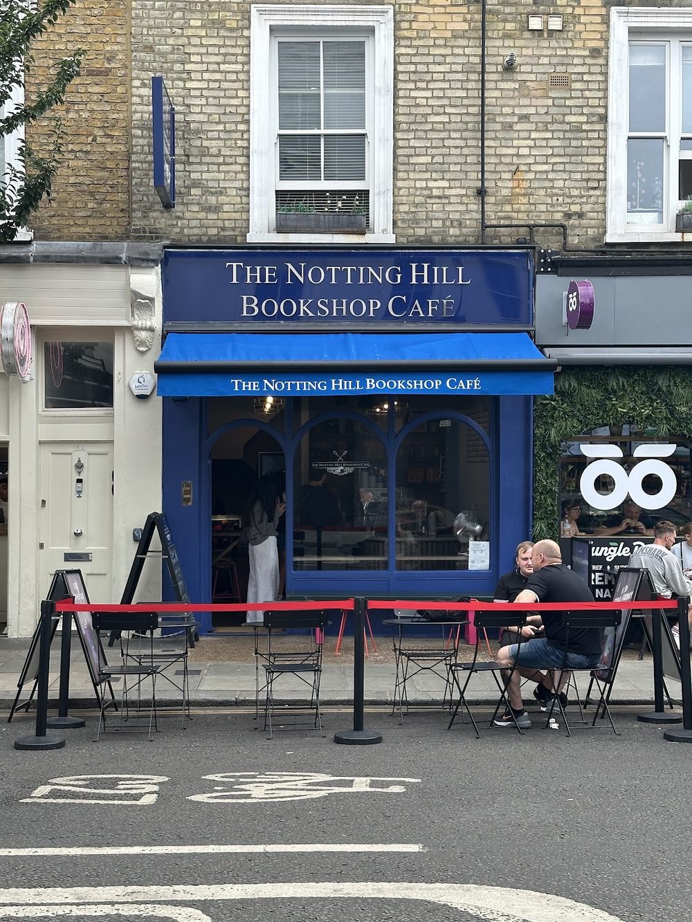 The blue shopfront of The Notting Hill Bookshop Café seen from across Blenheim Crescent, with red pavement rails and outdoor seating