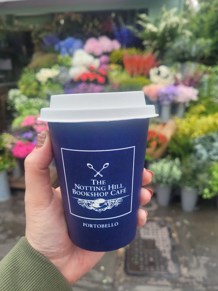 A hand holding a navy takeaway cup in front of a flower stall, cropped to remove lettering